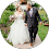 A person in a formal suit walks with another in a white dress, outside on a garden path, smiling and holding hands.