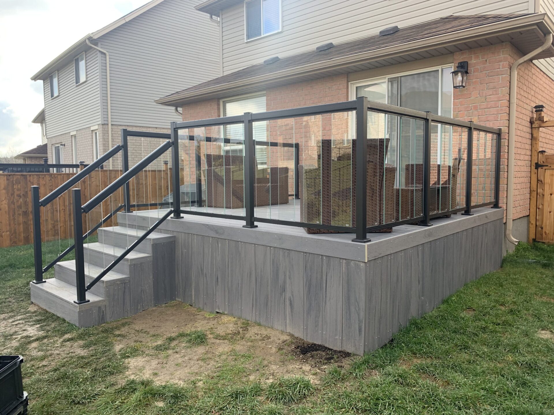 A house with a raised wooden deck featuring glass railings, wooden stairs, and outdoor furniture. Adjacent houses and a wooden fence are visible.