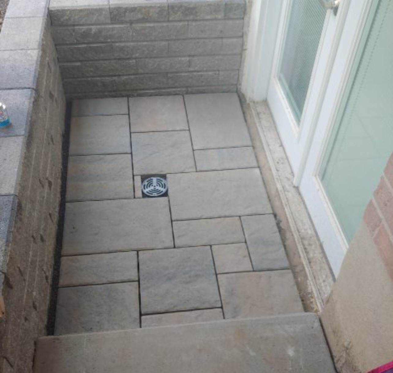 An outdoor area with stone tiles, a drain, and stairs leading to a white glass door, surrounded by grey brick walls.