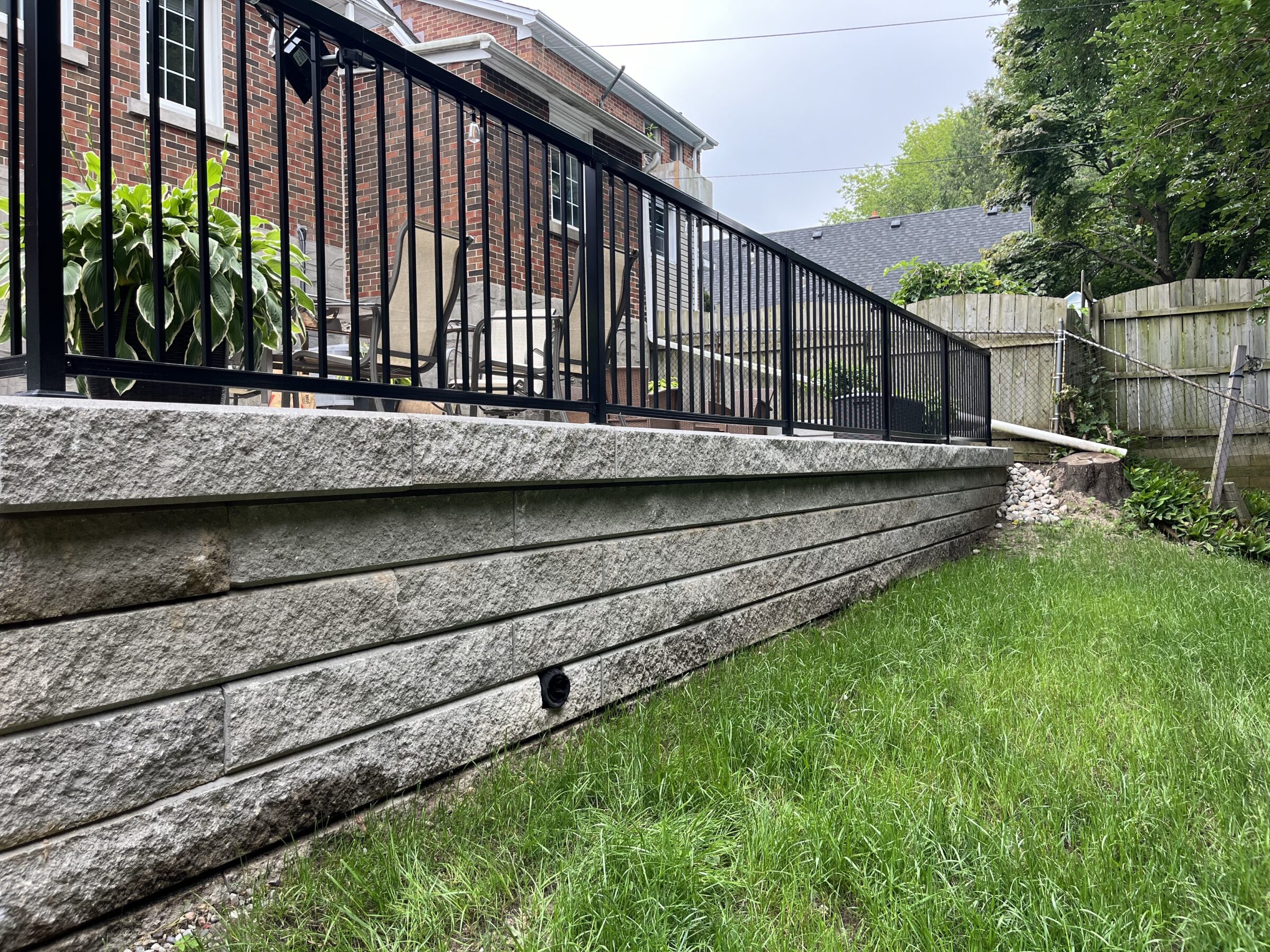 A backyard with a stone retaining wall, metal railing, lush green plants, and a brick house in the background under a cloudy sky.