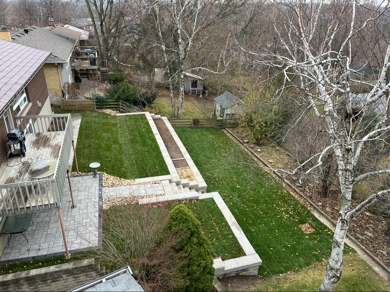 Aerial view of a backyard with a deck, stairs, manicured lawn, garden beds, mature trees, and adjacent houses in a suburban neighborhood.