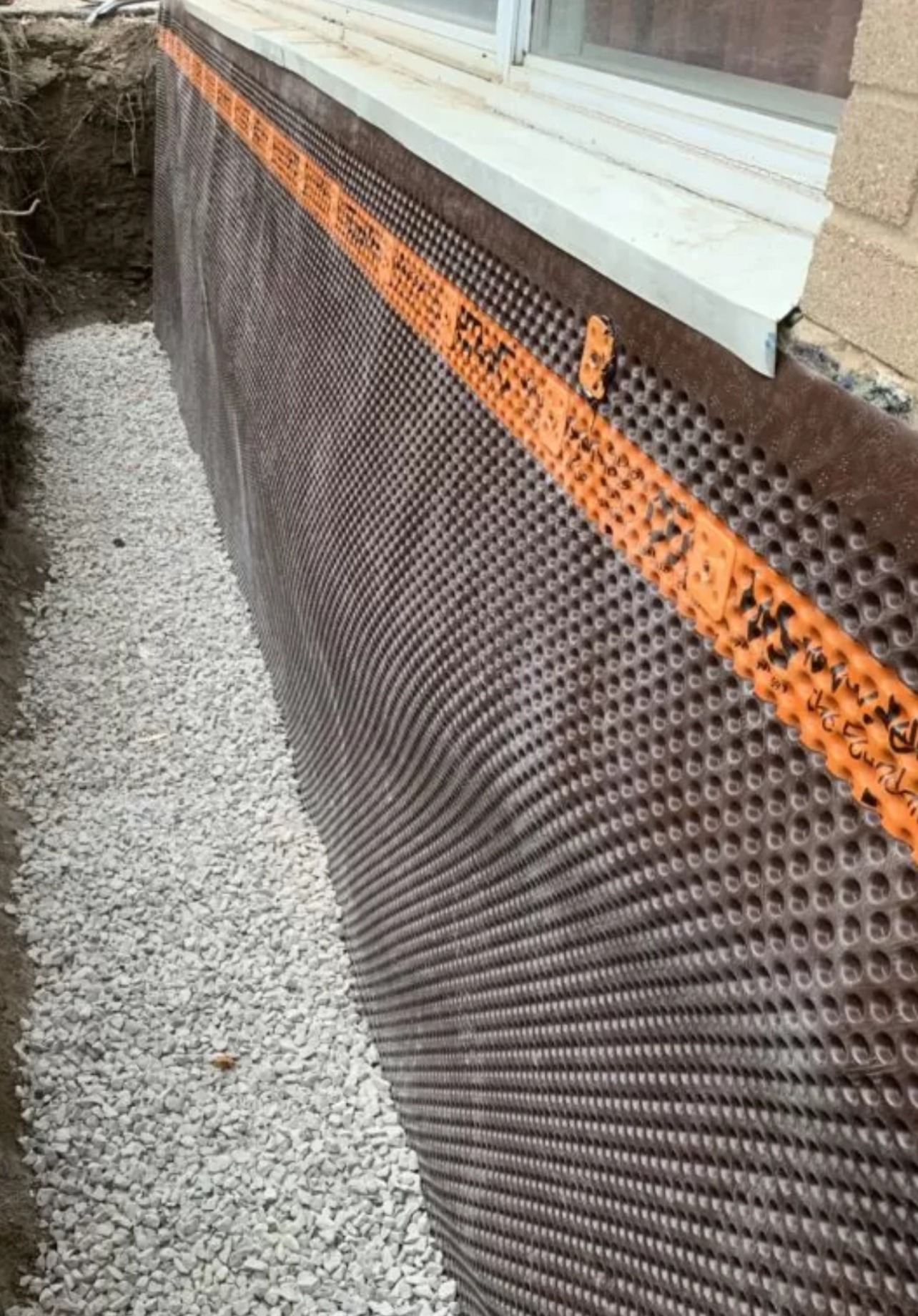 A construction site shows a newly installed waterproofing membrane on a building foundation with gravel at its base and an adjacent window.