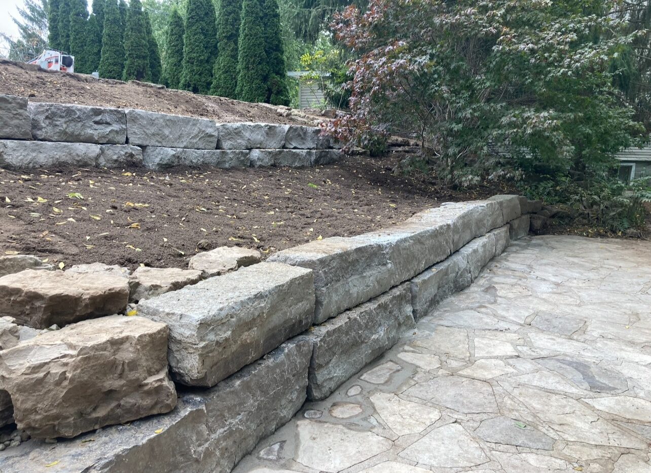 Stone retaining walls hold up a raised garden bed with freshly turned soil, surrounded by evergreen trees and bushes, next to a stone patio.