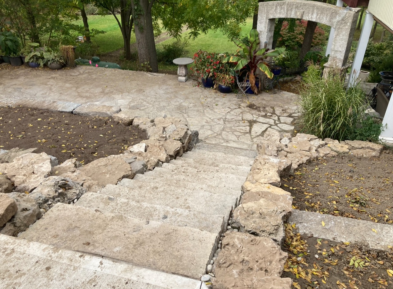 Stone steps lead down to a garden with lush greenery, potted plants, and a stone archway, bordered by trees in the background.