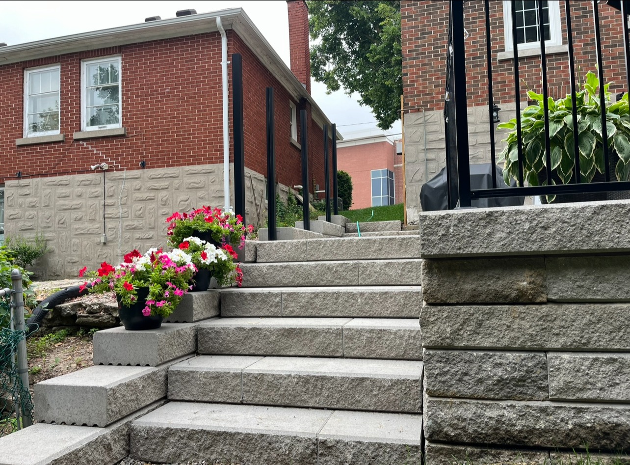 Concrete steps with potted flowers lead to brick buildings, surrounded by greenery. A black metal railing is partially visible on the right.