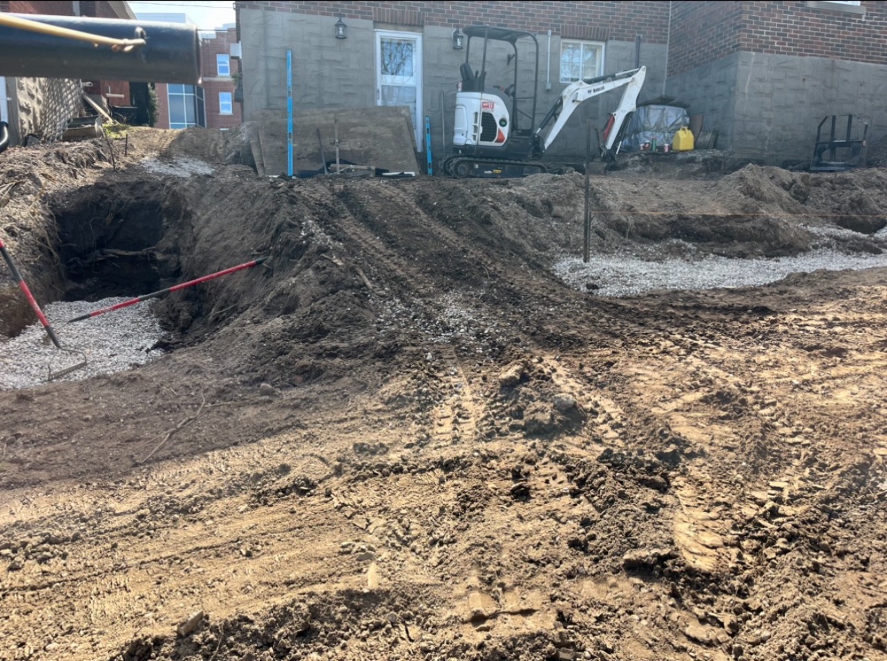 Construction site with heavy machinery, earth dug up, and gravel spread at the foundation of a brick building in a residential area.