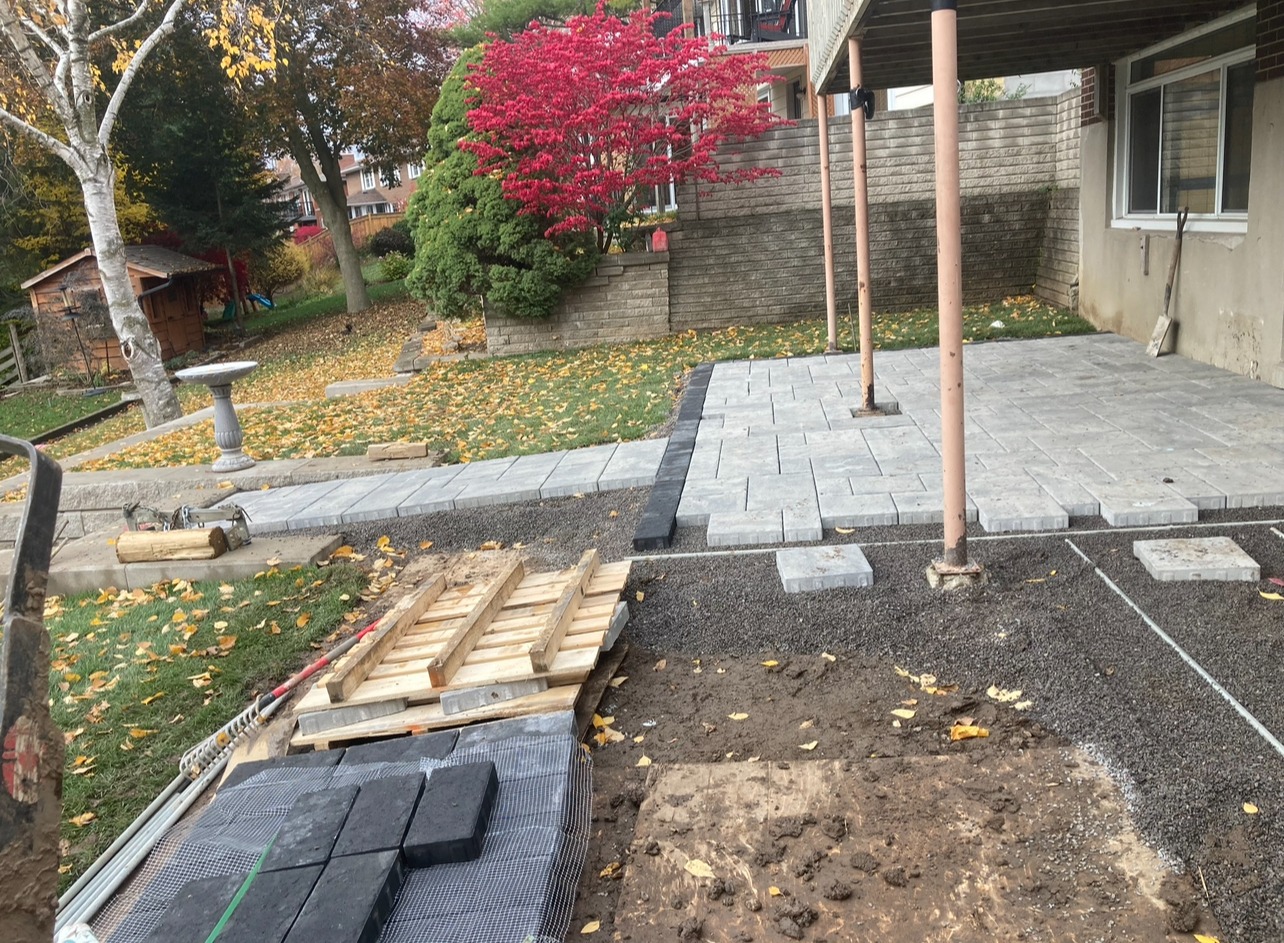 A newly constructed patio under a balcony, with a concrete walkway, colorful foliage, and a small garden shed in the backyard. No people visible.