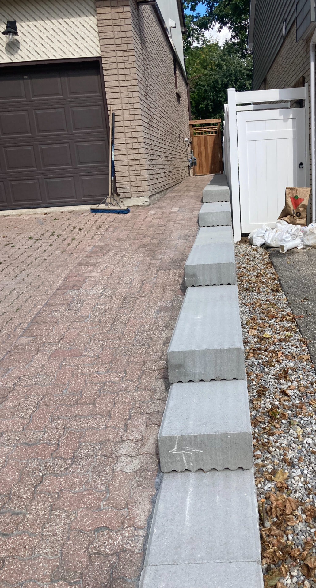 A residential driveway shows a stepped concrete border, a closed brown garage door, a nearby broom, and a white fence leading to a backyard.