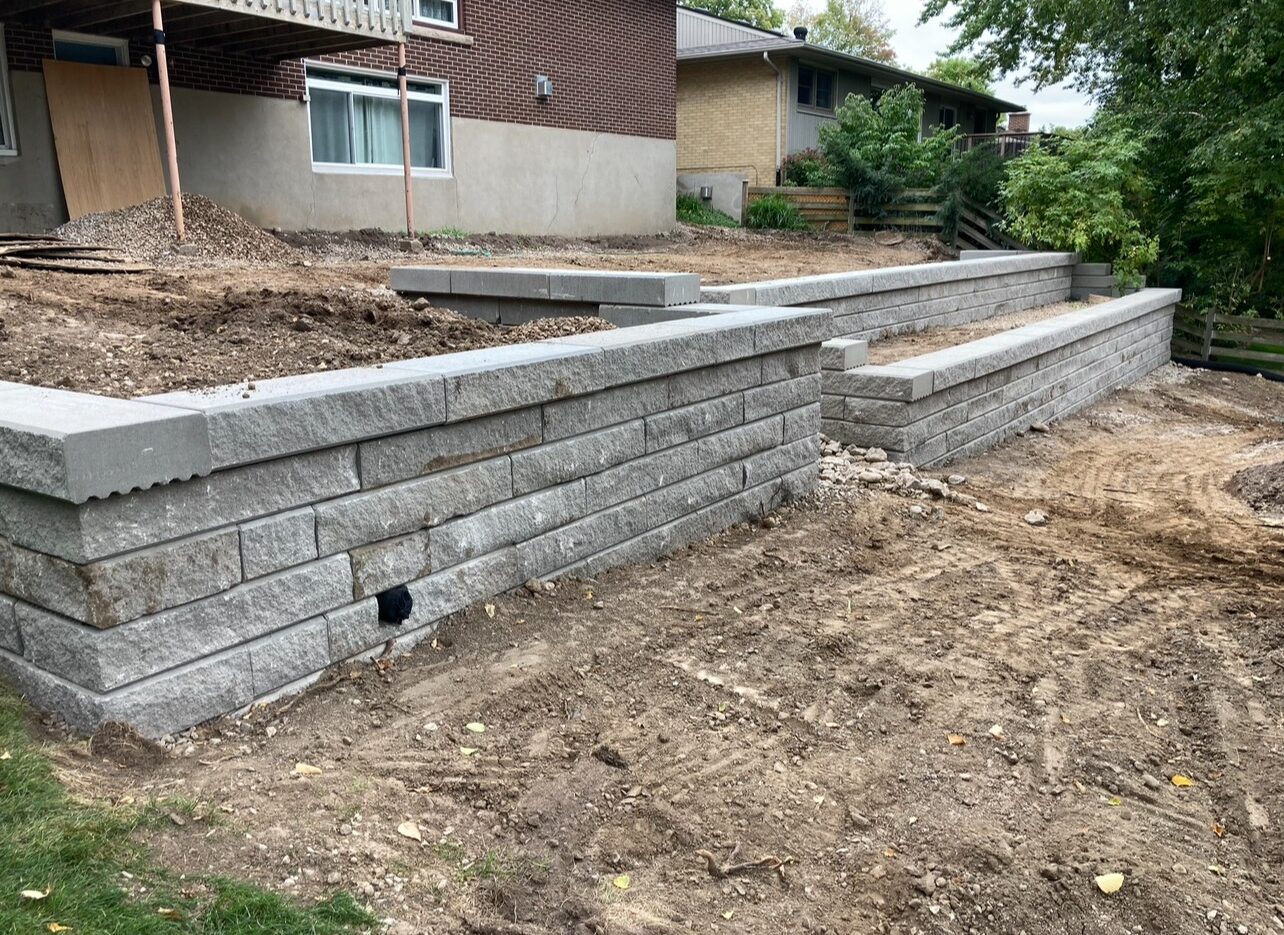 A newly constructed retaining wall in a residential backyard with exposed construction materials and soil, adjacent to a brick house.