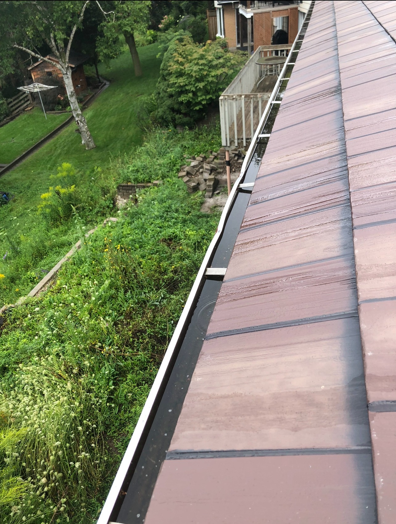 A view of a rooftop and gutter overlooking a lush green garden with a wooden deck, a tree, and a small shed.