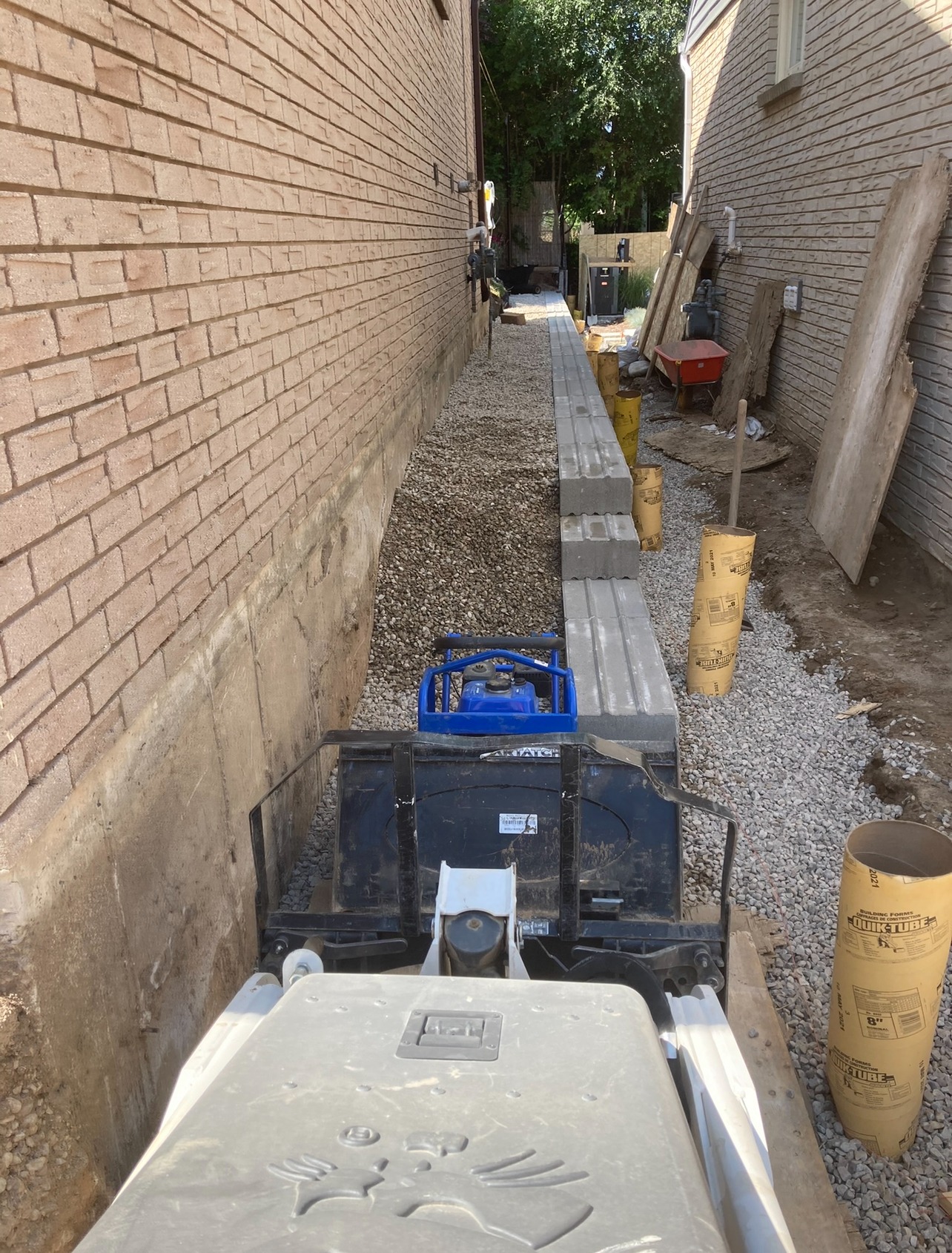 Two brick buildings with a narrow alleyway between them, under construction. Equipment and materials are placed along the gravel path, with one person visible.