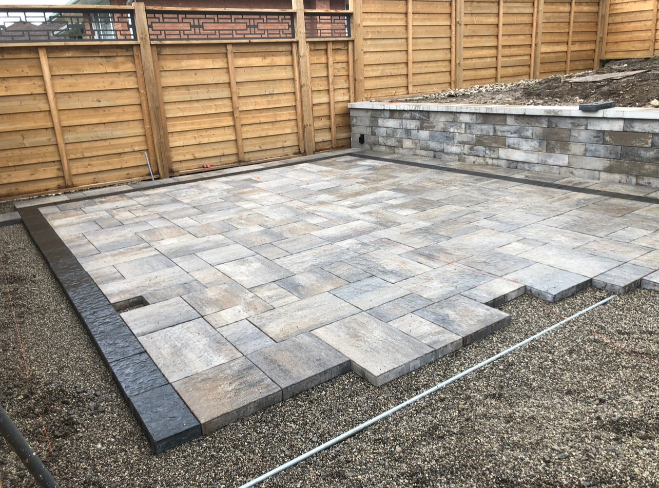 The image shows a newly paved patio area with grey bricks, wooden fence, and a short stone retaining wall, under construction. No landmarks present.