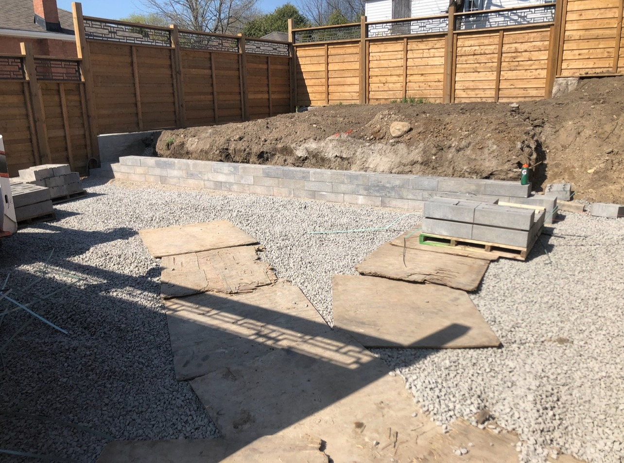 A construction site with gravel, wooden boards, a retaining wall, and a person working. A wooden fence with lattice tops encloses the area.