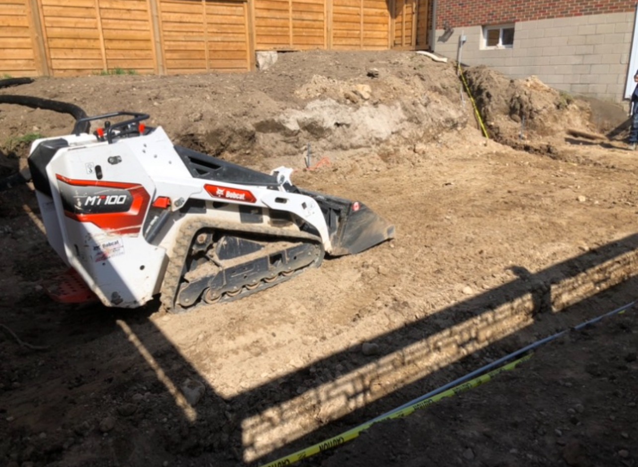 A construction site with a Bobcat MT100 loader, dirt, a wooden fence, brick wall, caution tape, and a person in the background.