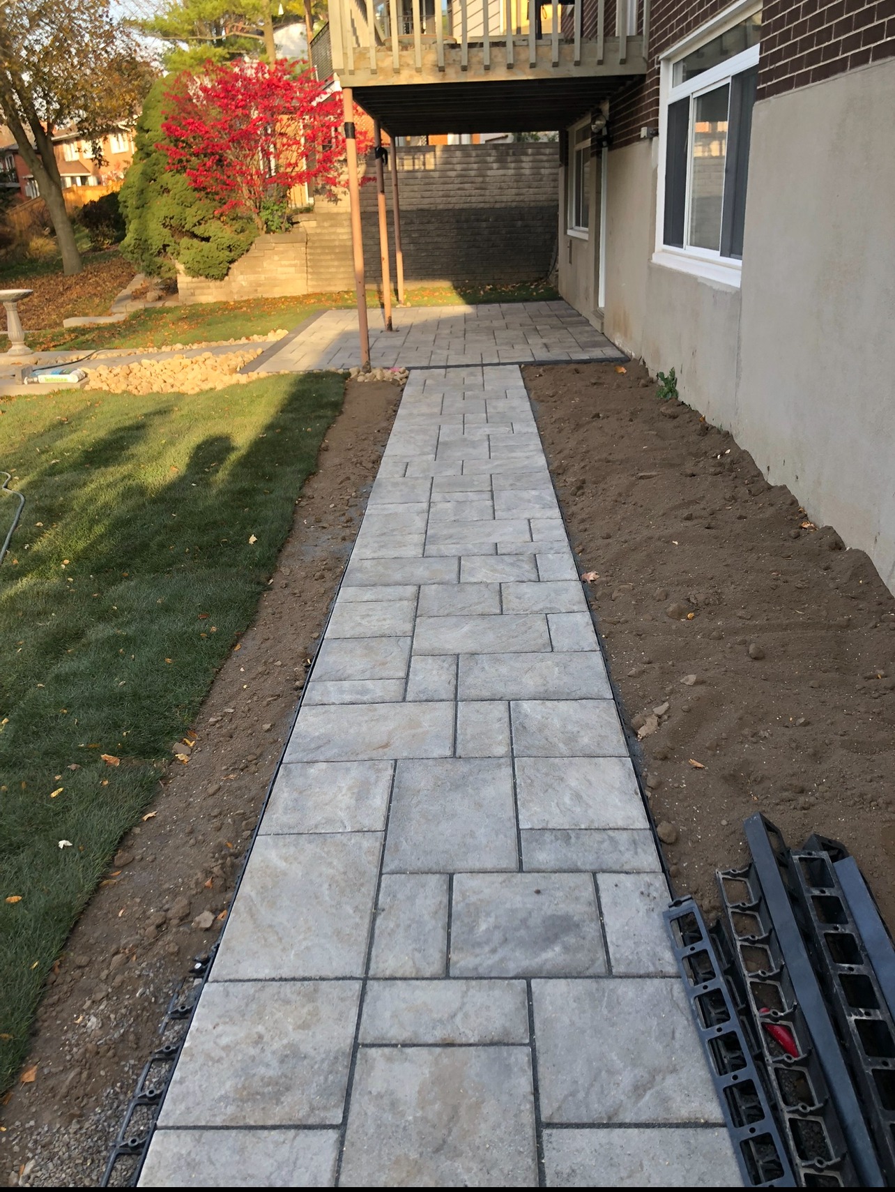 A stone pathway leads to a house's back deck with autumn trees and manicured lawn in the background.