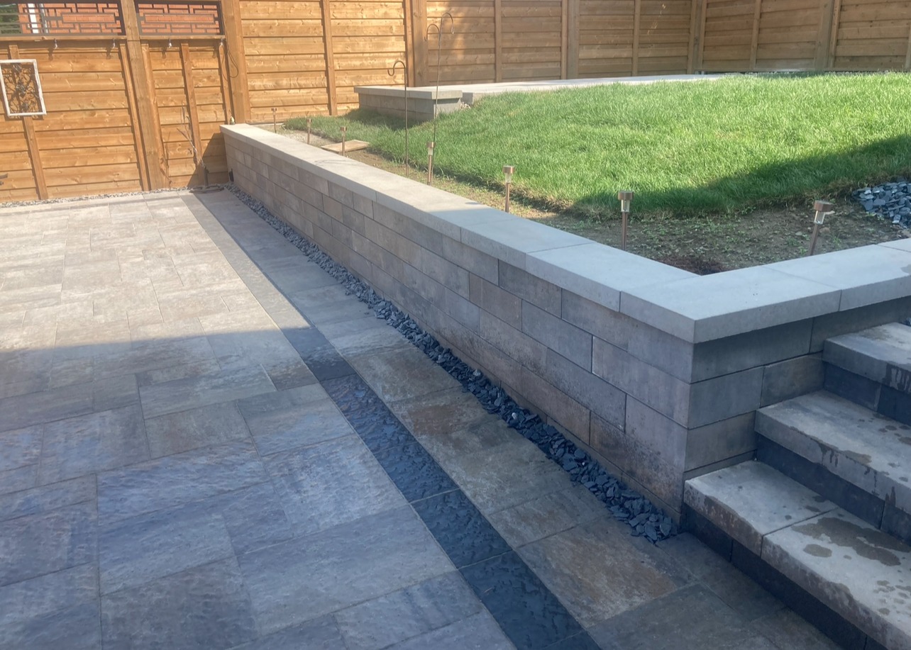 This image shows a modern backyard with stone tiles, a small elevated grassy area, and wooden fences. There are steps leading up to the grass.