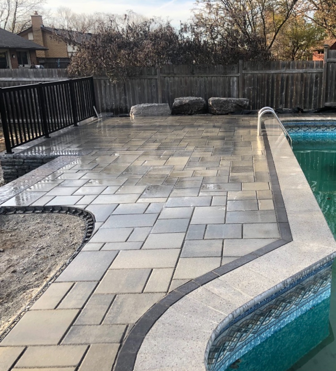 A backyard features a newly paved patio surrounding a swimming pool, bordered by a wooden fence with bare trees and houses in the background.