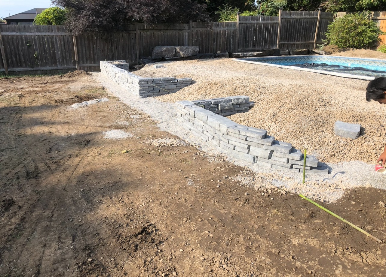 A backyard with a partially built stone retaining wall, dirt, gravel, a pool, and a person inspecting measurements near the construction.