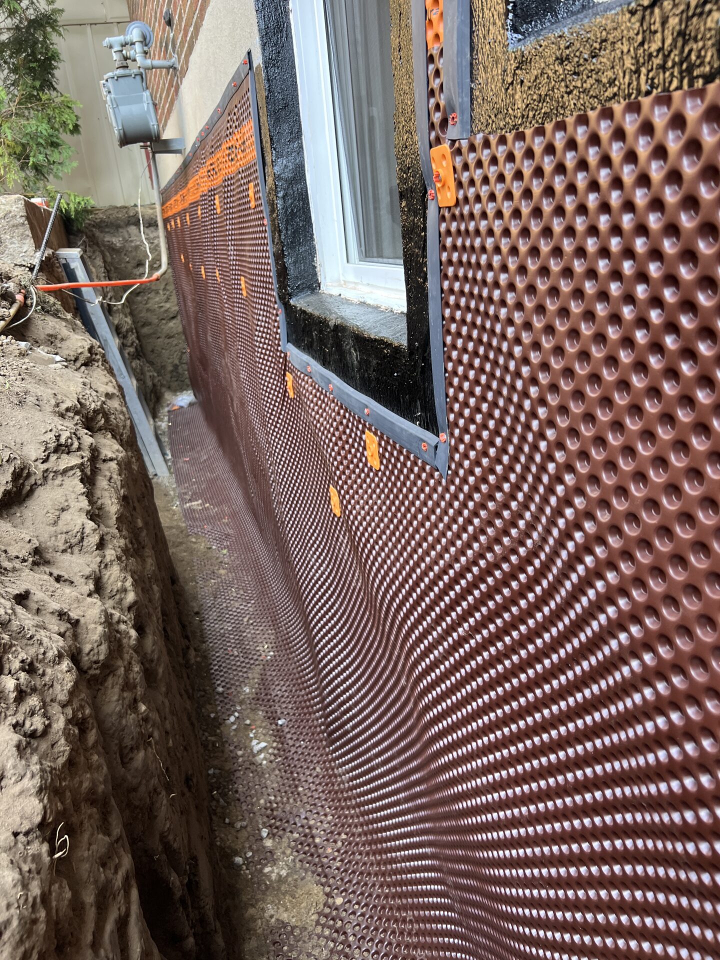 A close-up of a building foundation with waterproofing installed, adjacent to a trenched area, utility meter visible, partially covered with soil.