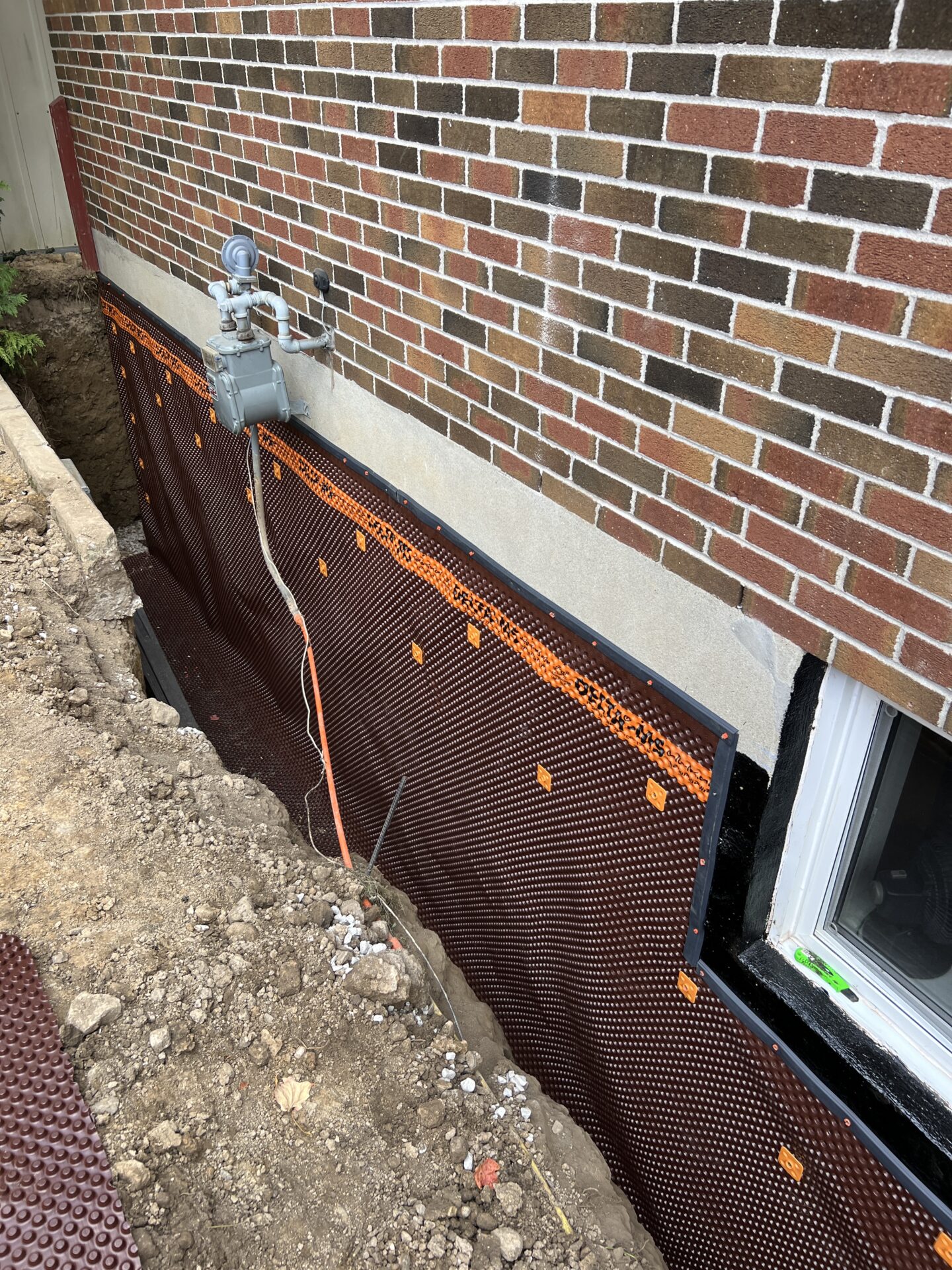 The image shows a section of a brick house foundation being waterproofed with a membrane. Nearby is an electrical box with pipes.