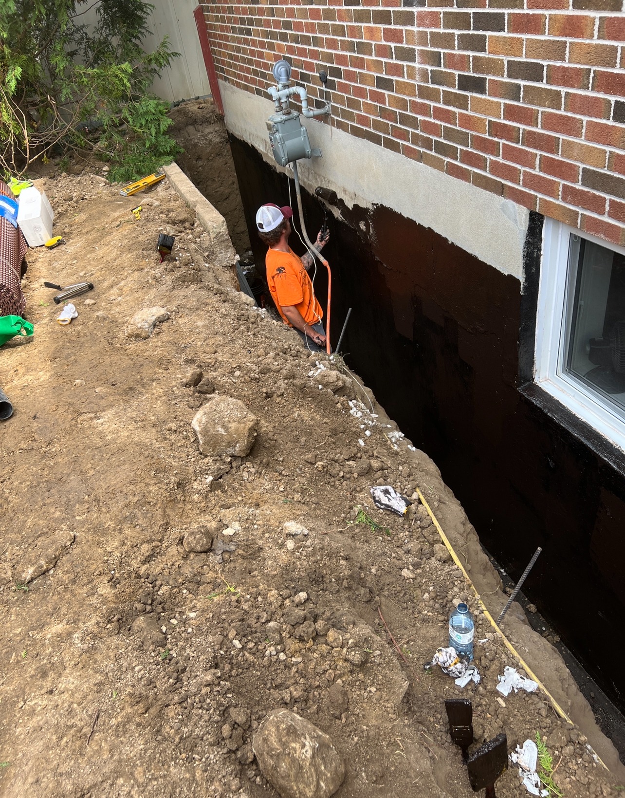 A person works on a home's exterior foundation in a dug-out trench, surrounded by tools, dirt, and rocks, near a brick wall.