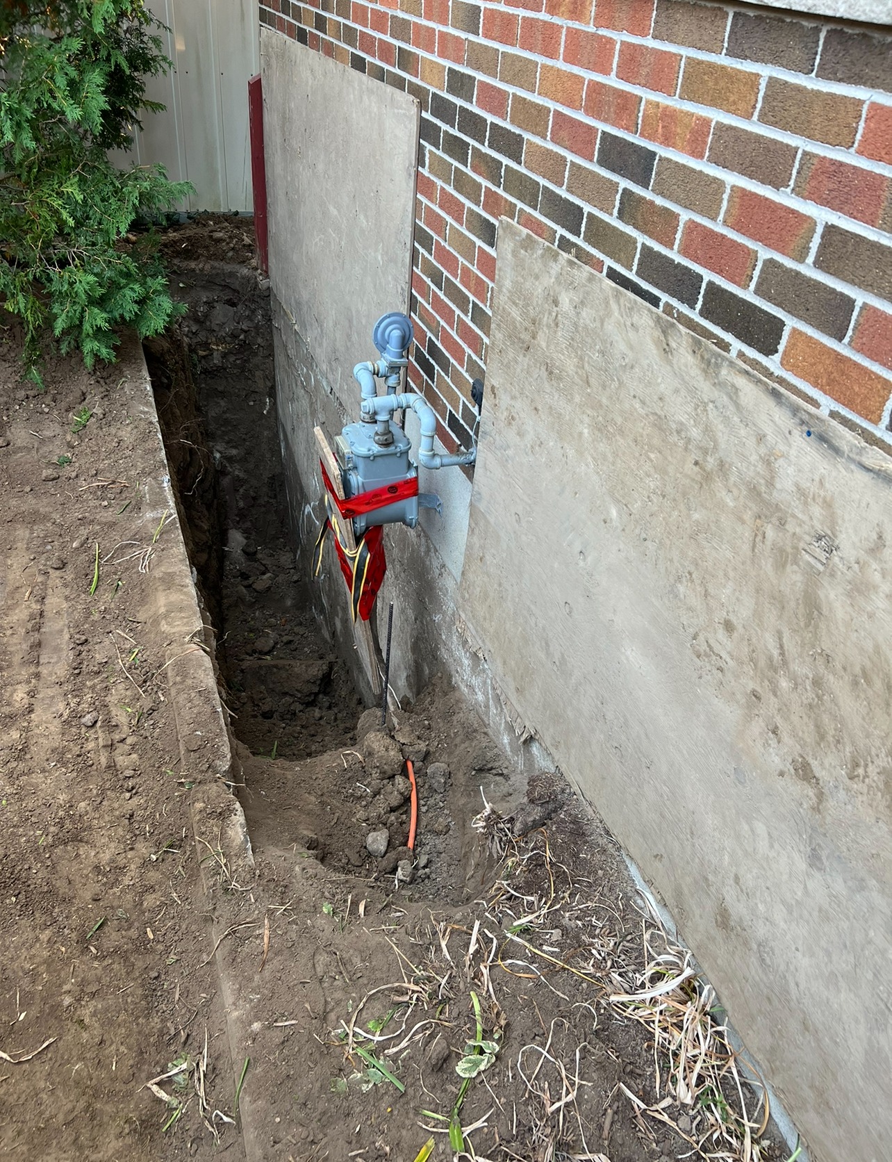 A trench beside a brick building under construction or repair, with visible piping and gas meter, and a tree near the corner.