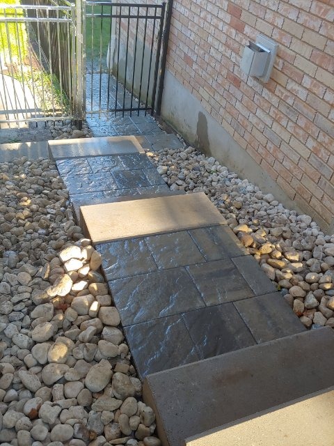 A wet stone pathway with rocks on the sides leads to a metal gate beside a brick wall, with sunlight casting shadows.