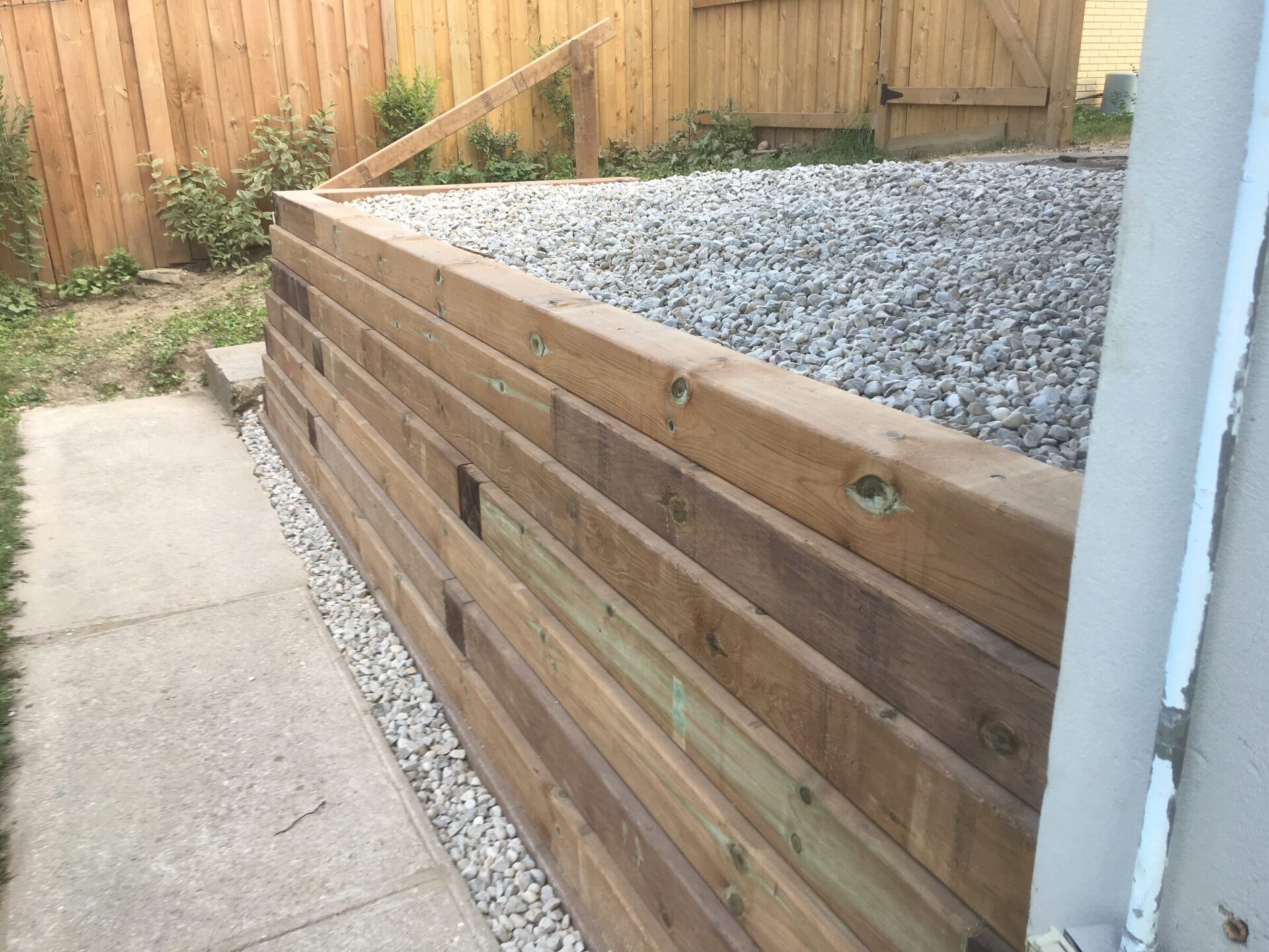 A backyard features a newly constructed wooden retaining wall filled with gravel, adjacent to a paved walkway and bordered by a wooden fence.