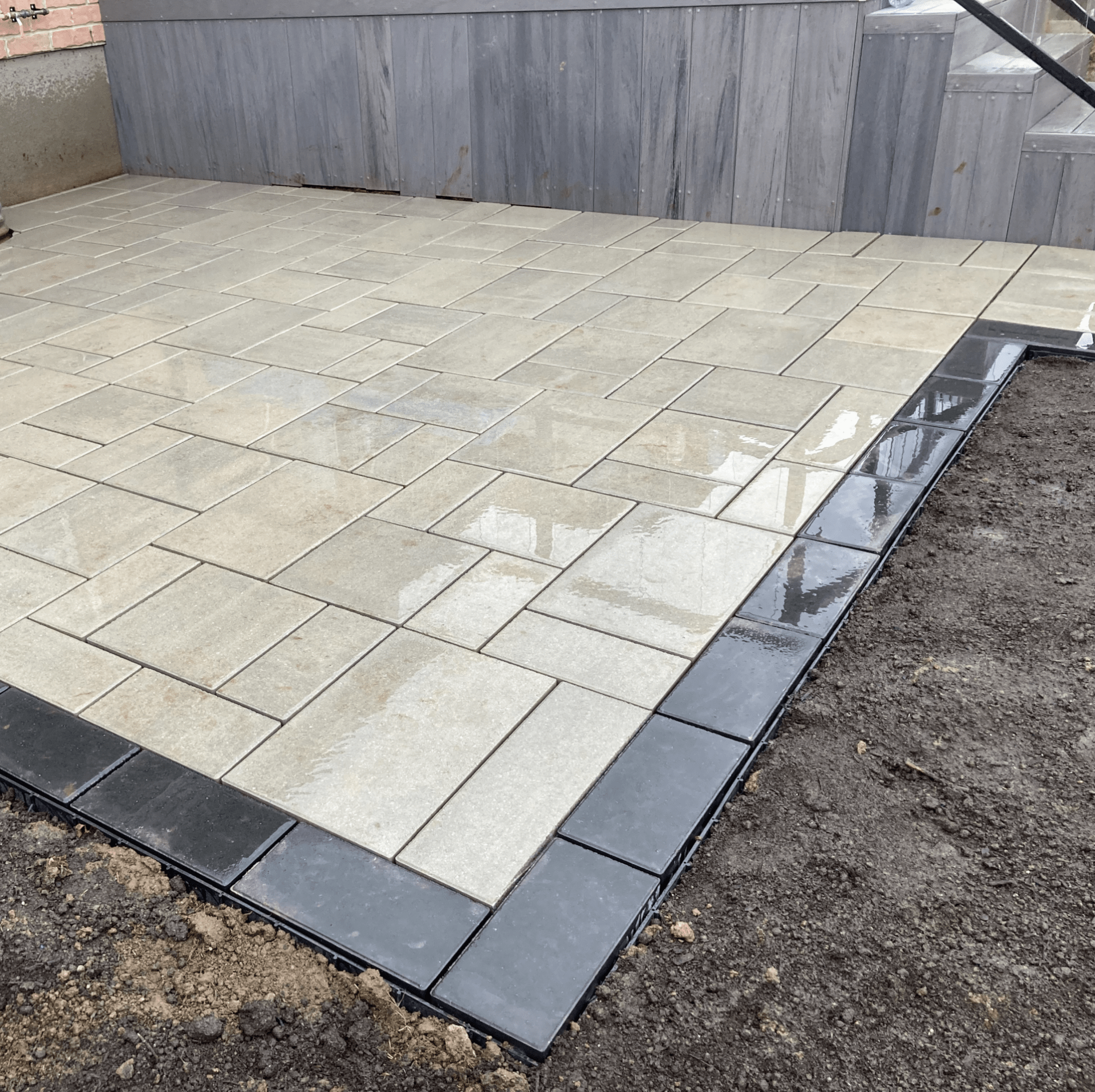 Freshly laid paving stones with a black border, adjacent to a modern wooden structure and surrounded by bare soil, showcasing recent construction.