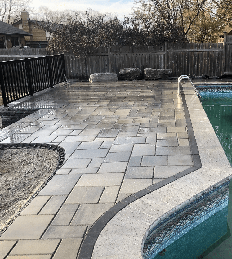 A backyard scene featuring a swimming pool with intricate stone pavers, a metal fence, and trees in the background, surrounded by a wooden fence.