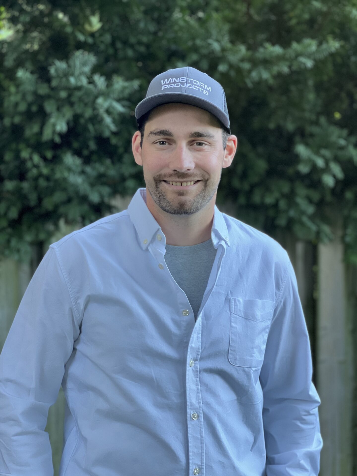 A person wearing a "WINSTORM PROJECTS" cap and light blue shirt stands outdoors with a background of leafy greenery.