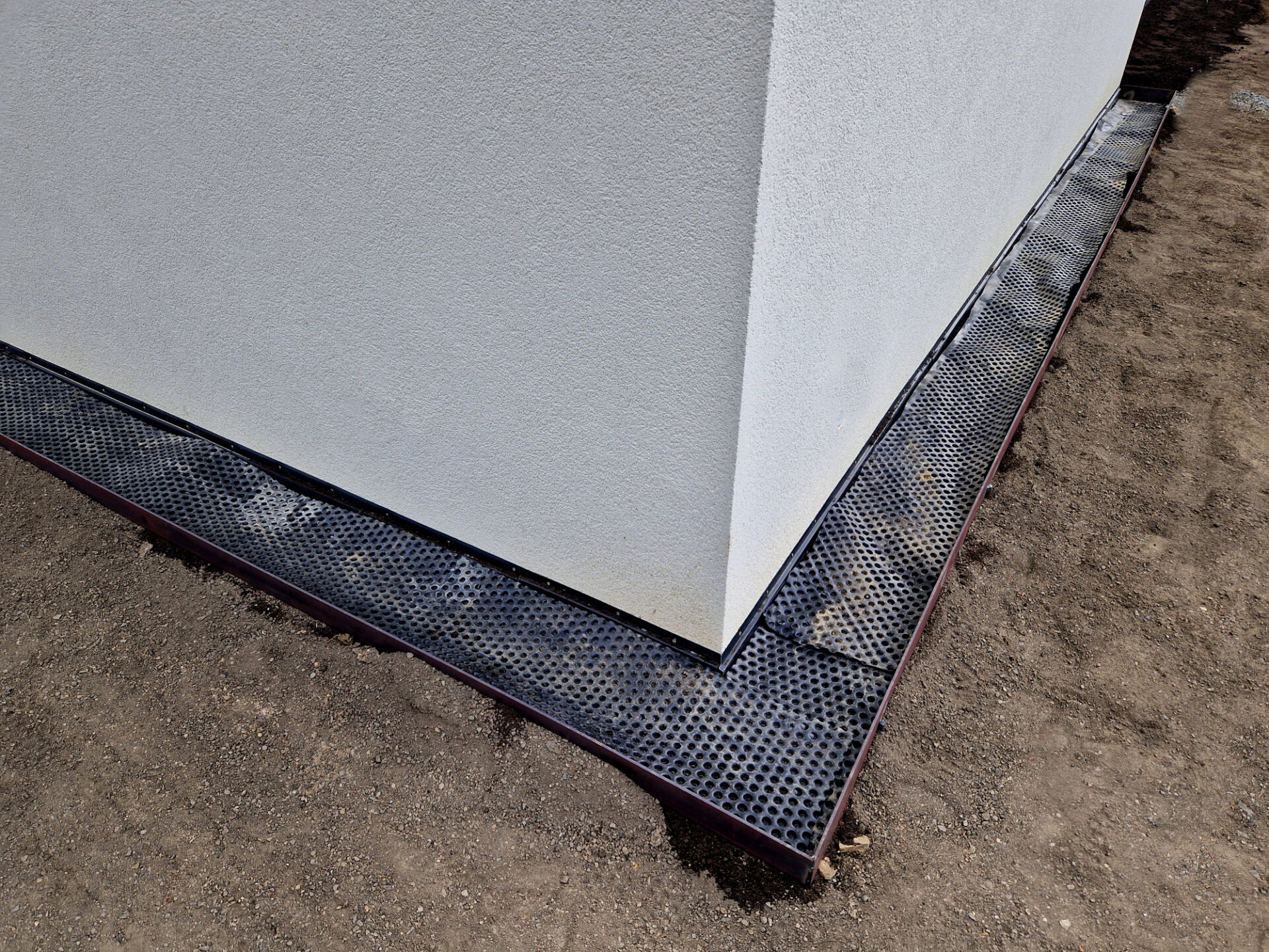 A close-up of a building's foundation corner with black perforated material and surrounding dirt, likely indicating construction or drainage installation work.