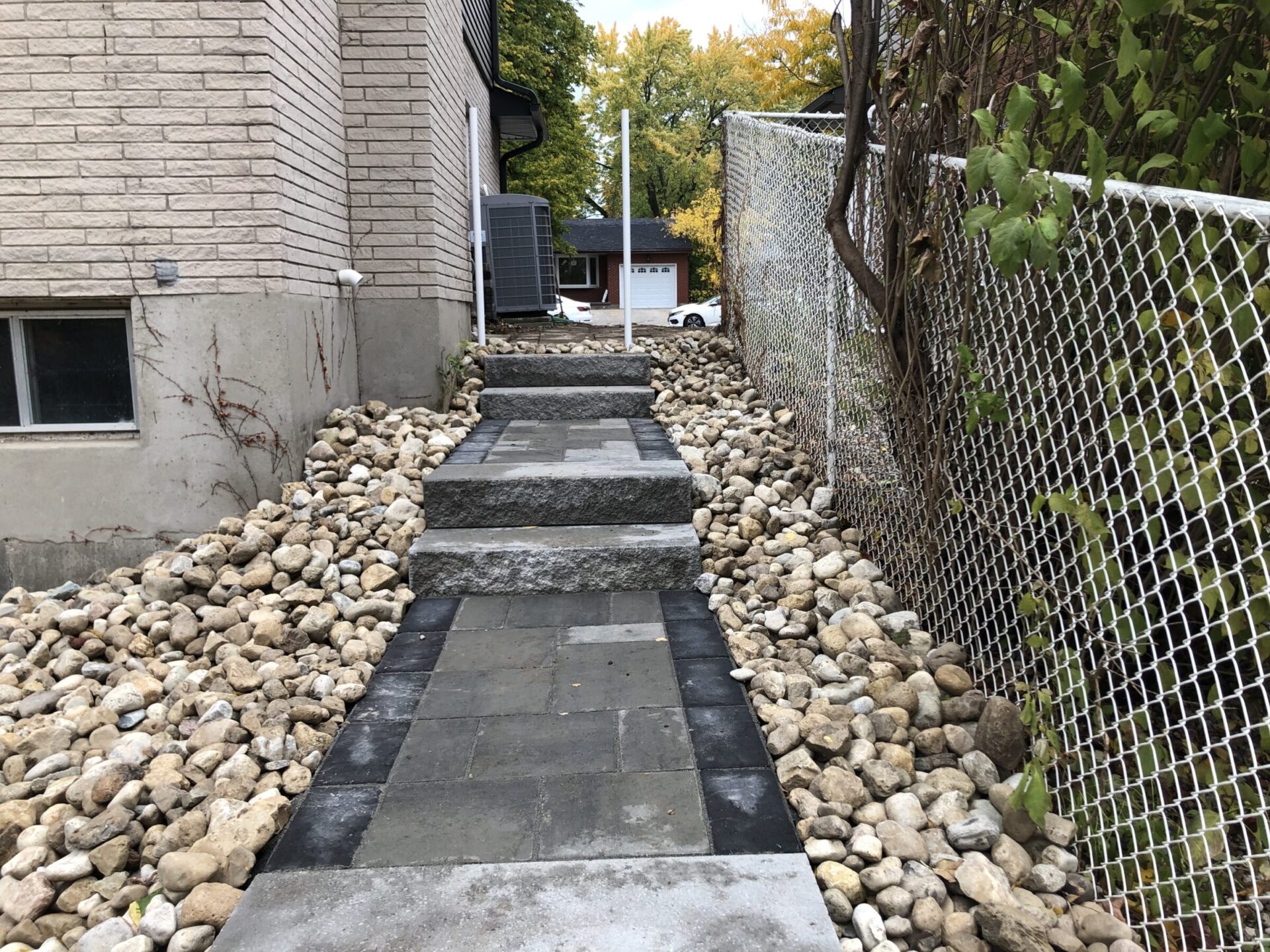 A paved walkway with stone steps lies between a house and a fenced area, leading to a driveway with a garage in the background.