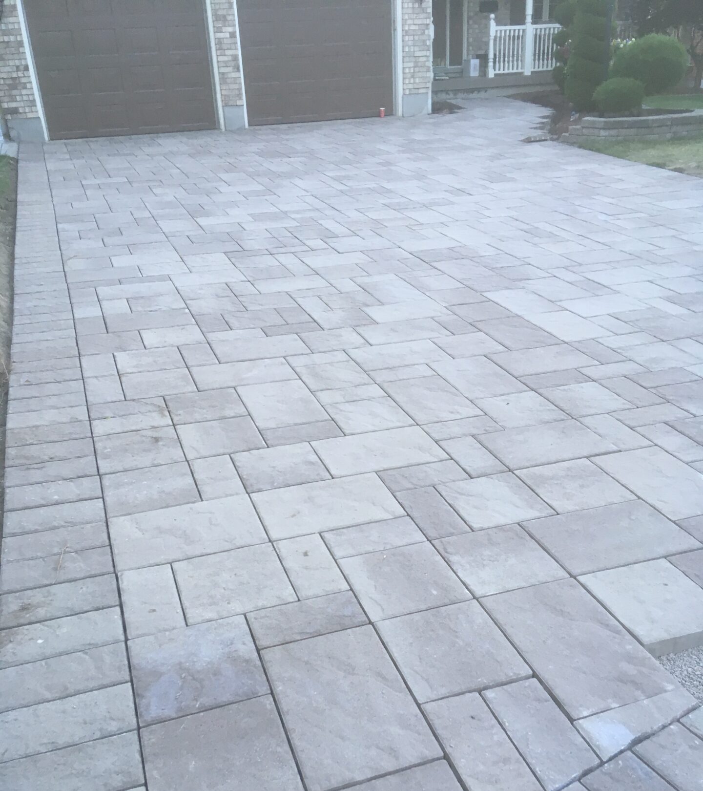 A freshly constructed paved driveway leads to a house with brown garage doors and a porch, featuring a well-maintained garden.