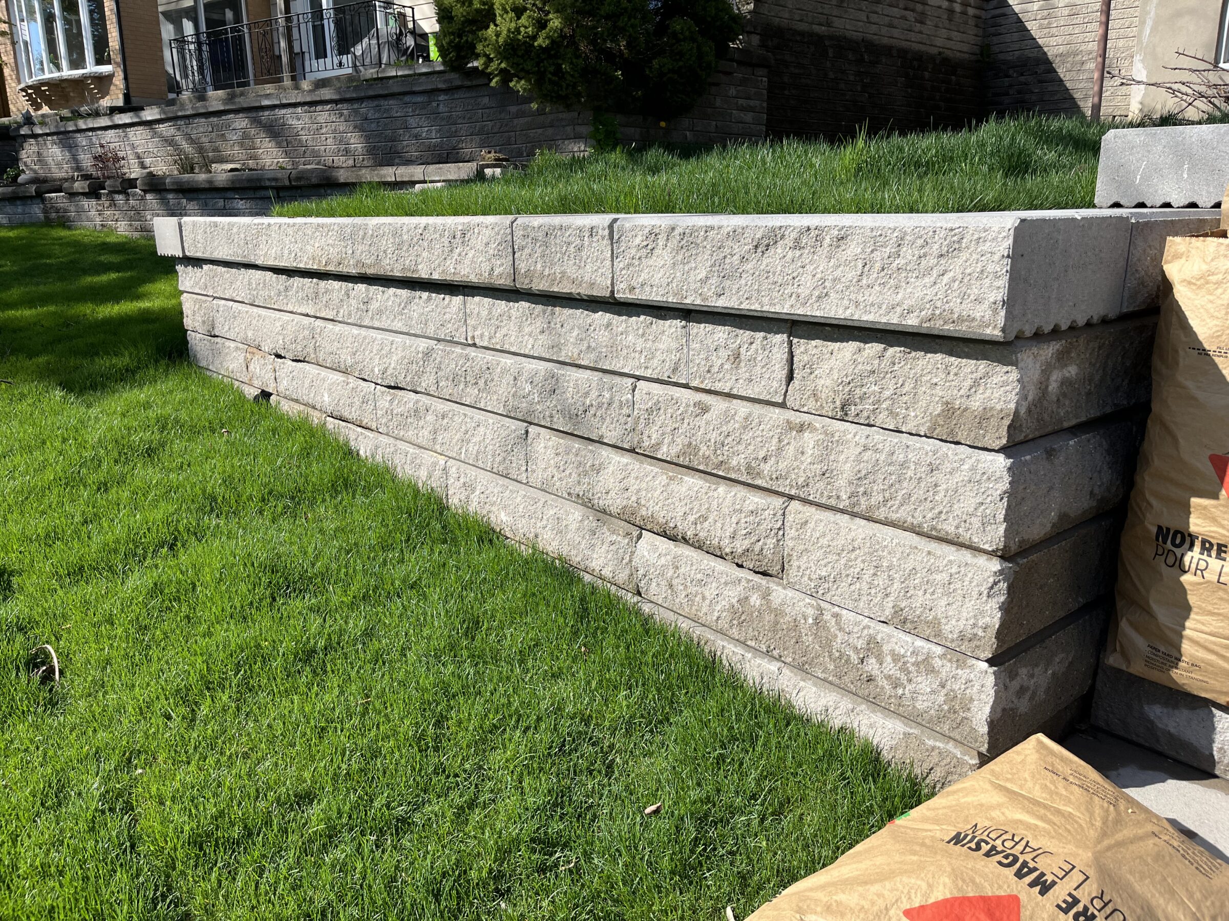 A neatly stacked stone retaining wall with lush green grass, adjacent to a house with red brick and a small balcony.