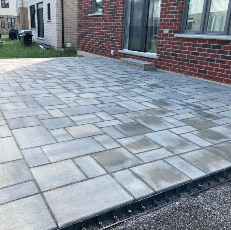 A newly paved patio with geometric stone tiles is adjacent to a red brick house in a residential neighborhood, featuring a green lawn.