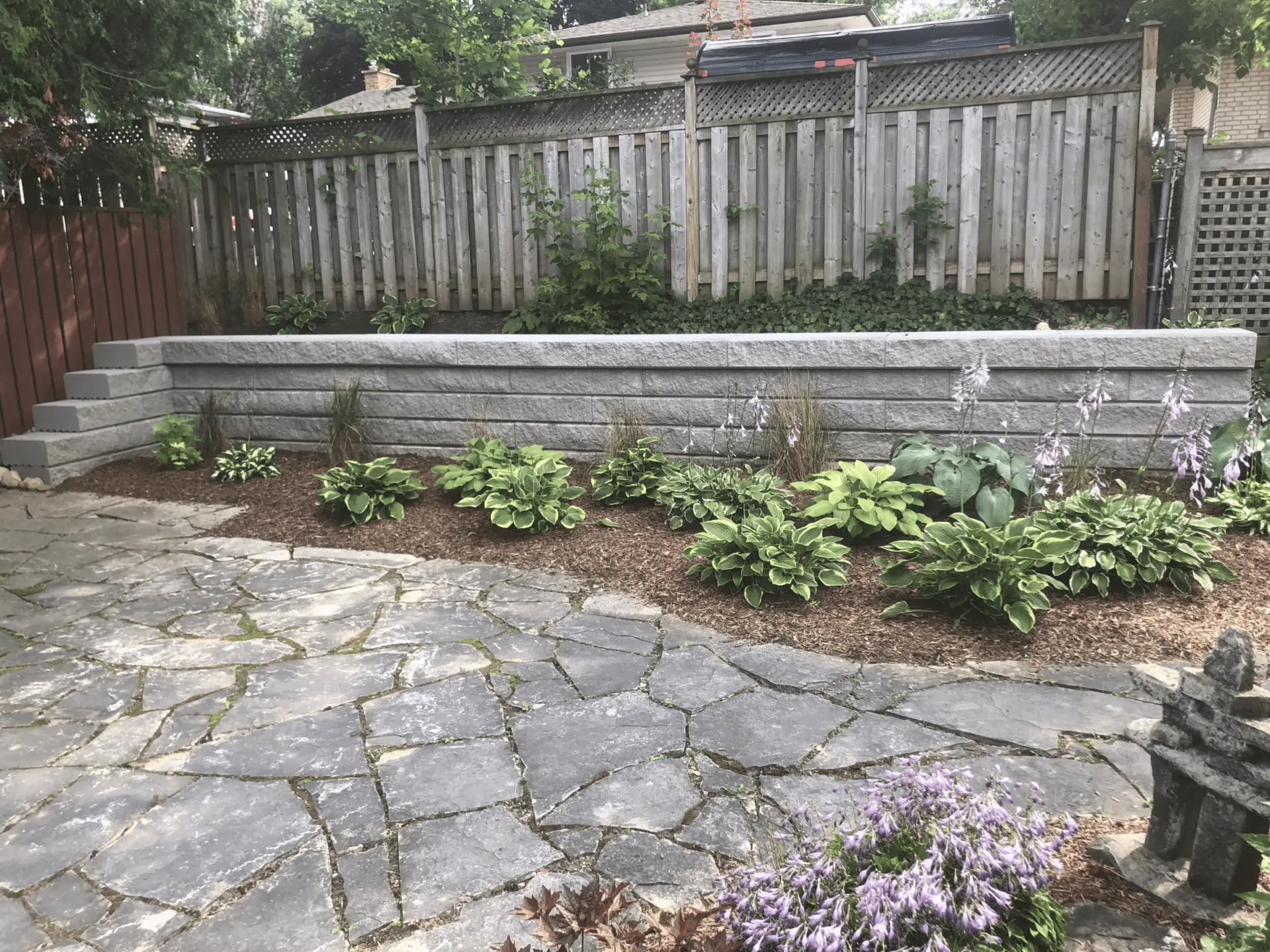 Stone patio with a raised garden bed, hostas, and ornamental plants near a wooden fence in a residential backyard setting.