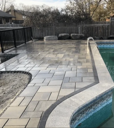 A neatly paved patio with a patterned tile design surrounds a swimming pool, enclosed by a wooden fence and black railing.