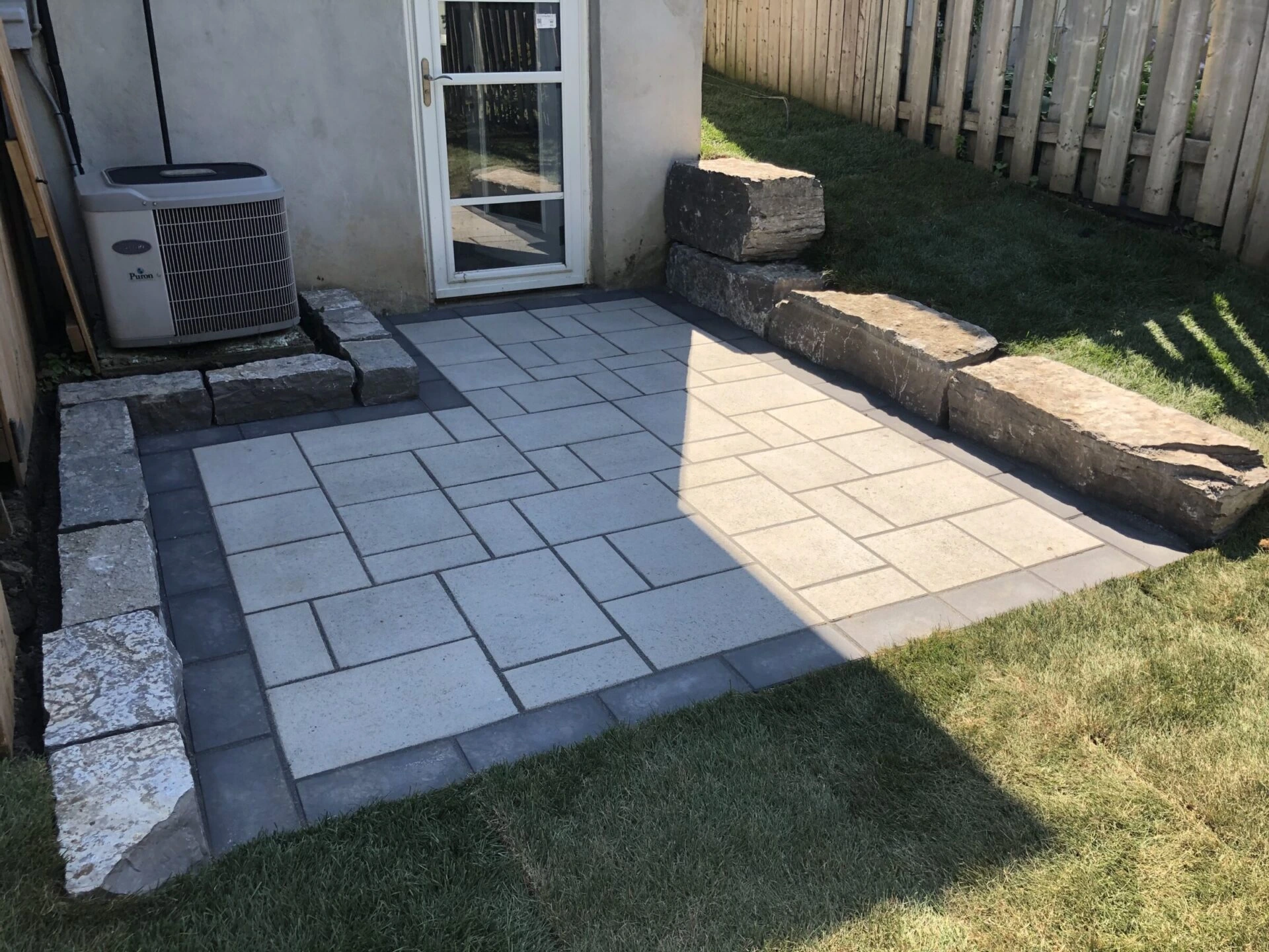 A small backyard patio features stone tiles and a retaining wall, surrounded by grass, near a house's back door and air conditioning unit.