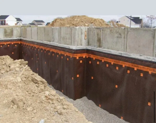 Construction site with exposed foundation and waterproofing material. Dirt piles surround the area. Nearby are suburban houses under a clear sky.