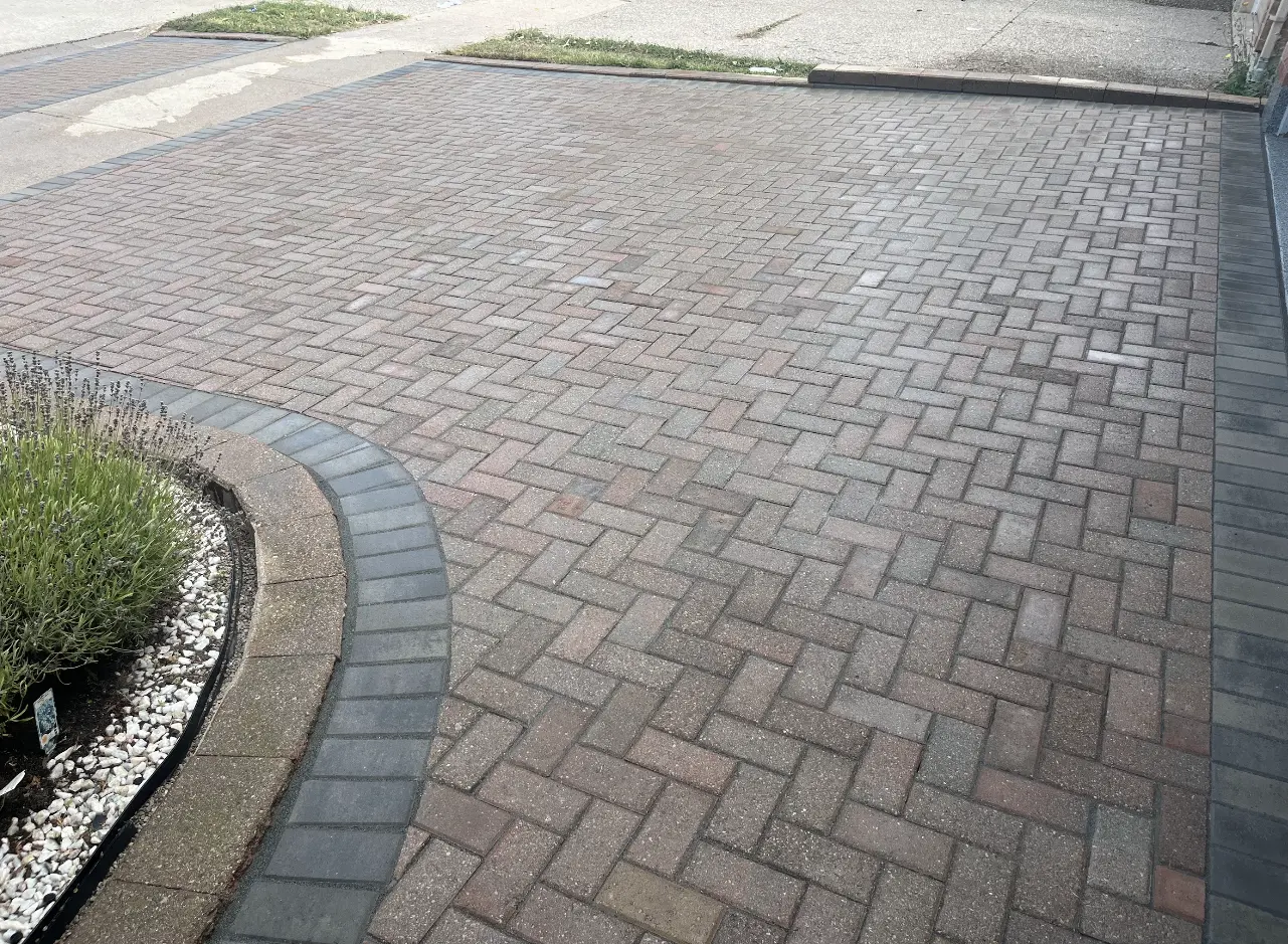 A paved brick driveway with a curved border frames a small garden bed filled with greenery and decorative white pebbles.