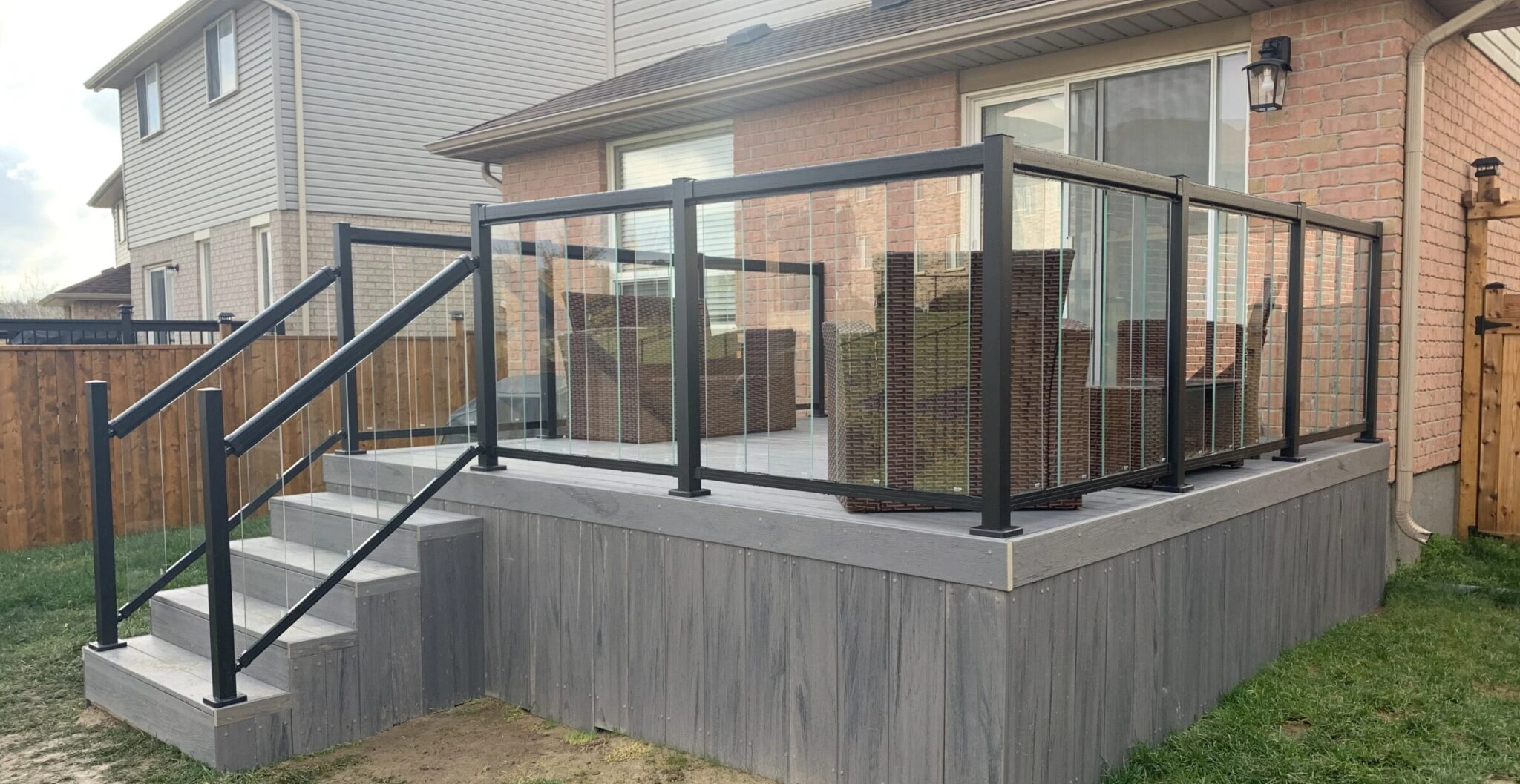 A modern backyard deck with transparent glass railings, wooden stairs, and wicker furniture, adjoining a brick house with large windows.
