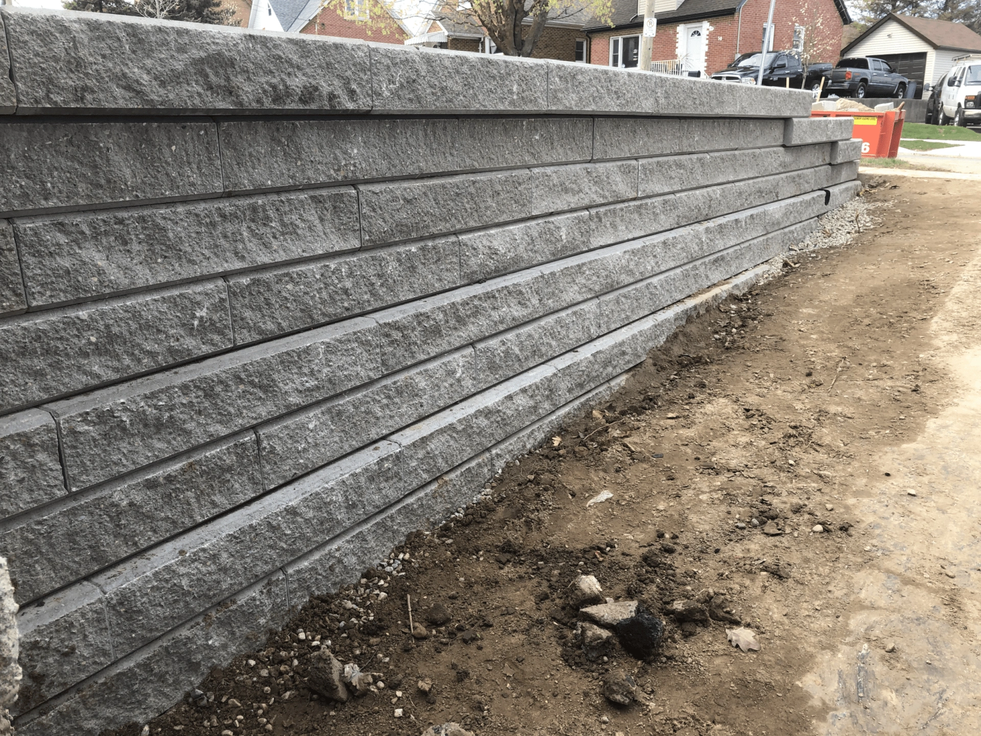 A newly constructed stone retaining wall lines a dirt pathway in a residential neighborhood, with parked vehicles and houses in the background.