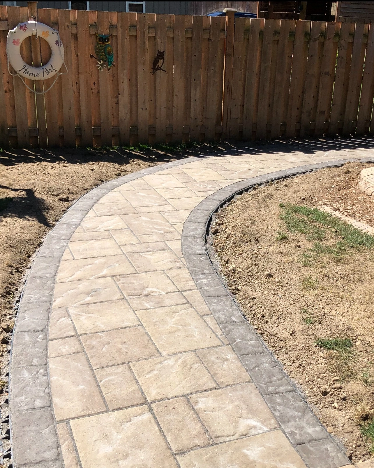 A curved stone pathway leads through a backyard with a wooden fence, adorned by metal owl decorations and a decorative life preserver.