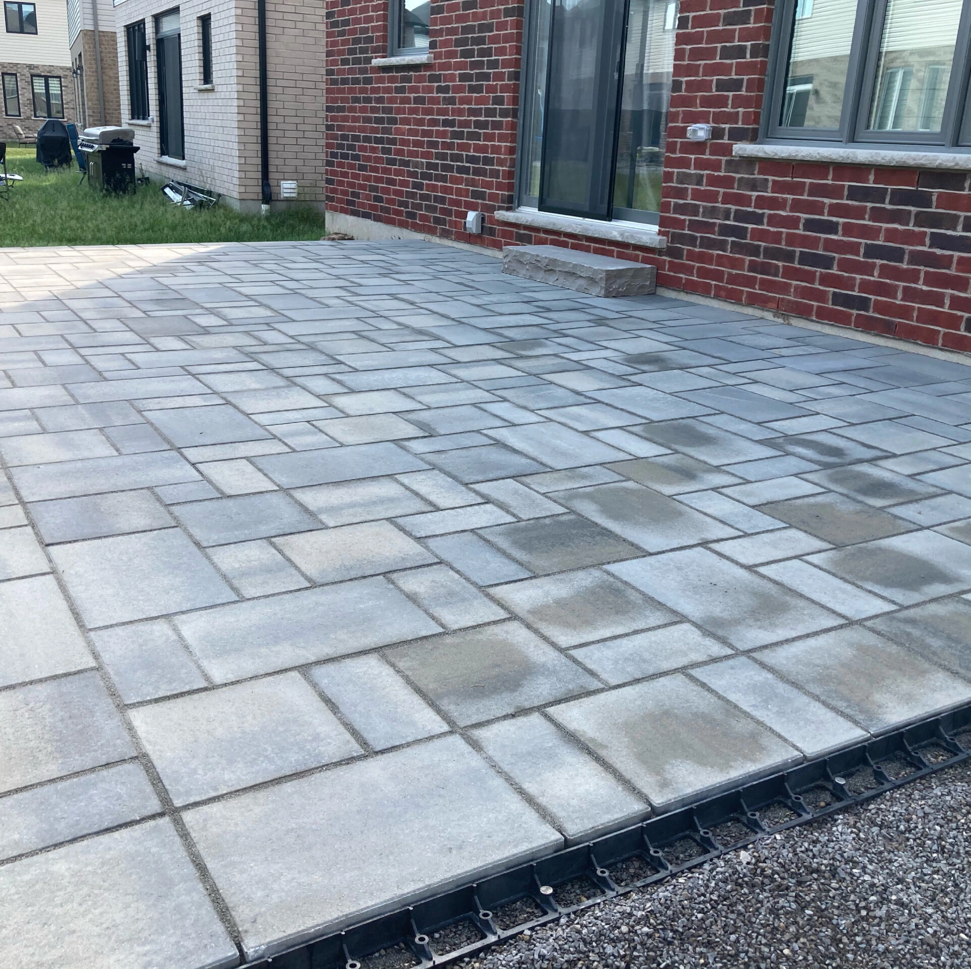 A newly laid stone patio with interlocking tiles next to a brick house, adjoining a small grassy backyard with nearby buildings.