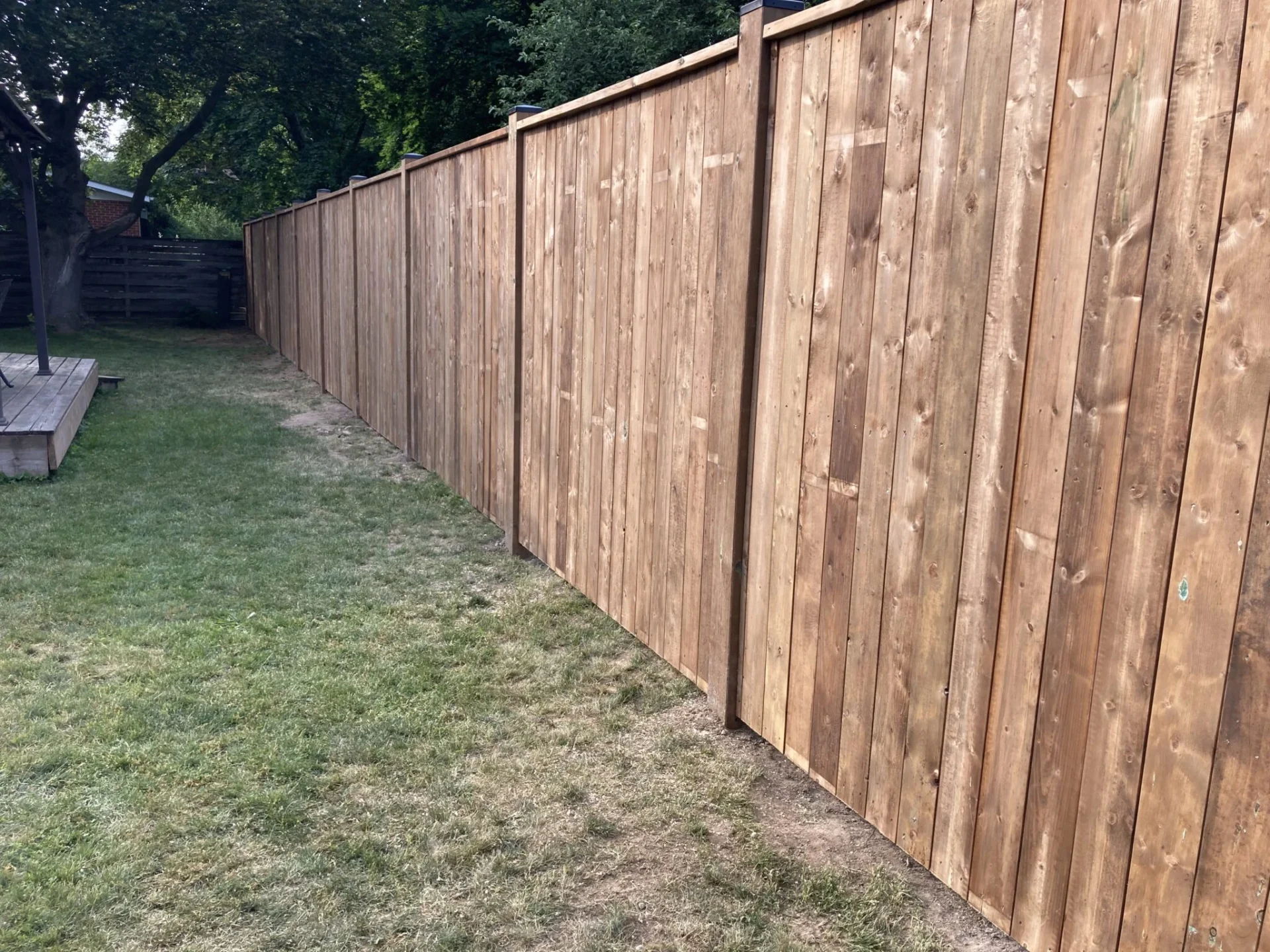 A wooden fence lines a backyard with a grassy lawn, adjacent to a small deck and surrounded by trees.