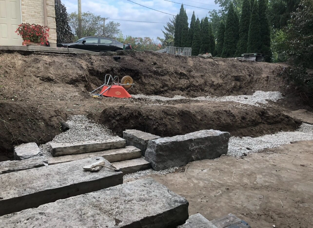 A construction site features a wheelbarrow, stone blocks, and a parked car amidst dug-up earth and gravel, with trees in the background.