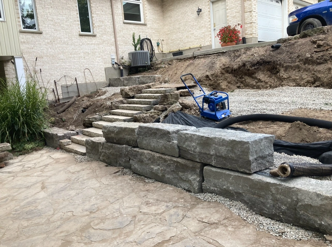 A backyard under construction with stone steps, large rocks, gravel, and a blue compactor. Brick house and plants in the background.