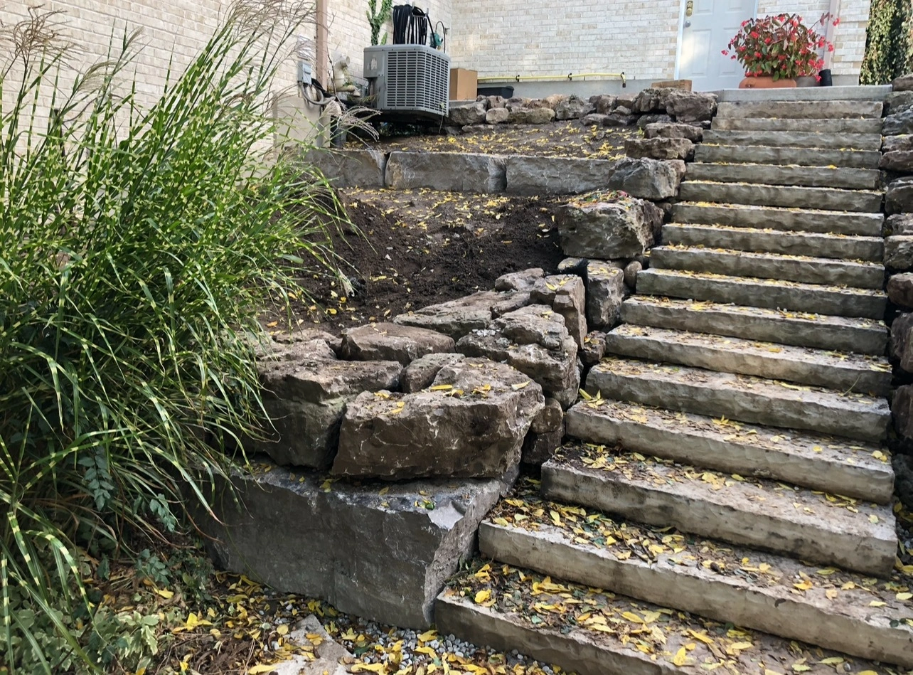 Stone steps covered with fallen leaves lead to a house. Lush greenery is on the left, with large rocks flanking the path.