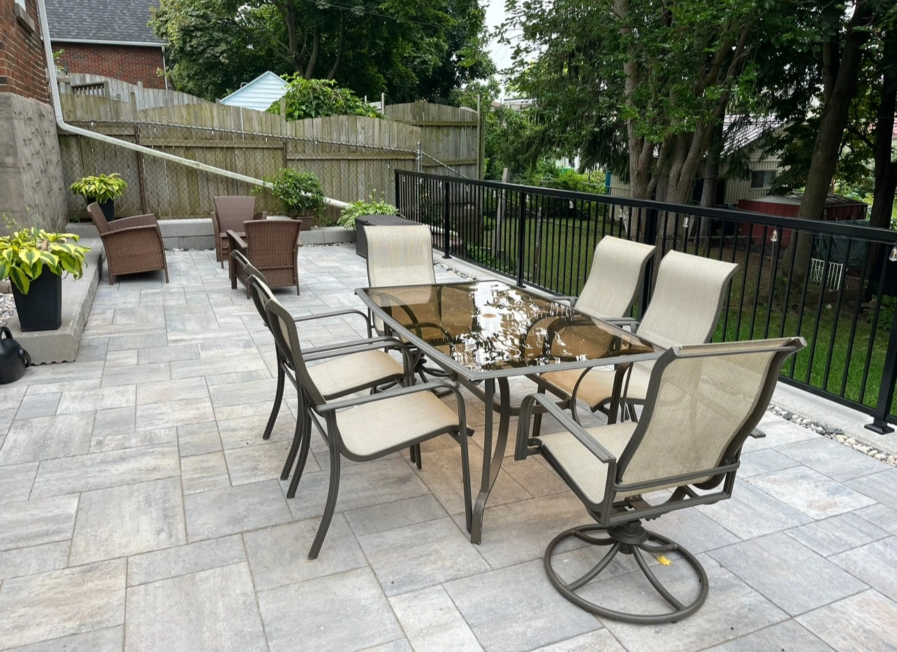 Outdoor patio with a glass-topped table, six chairs, wicker seating, and potted plants, surrounded by a wooden fence and greenery.